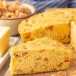 Thick slice of crackling bread on a wooden board with butter, pork cracklings, and a skillet of cornbread in the background