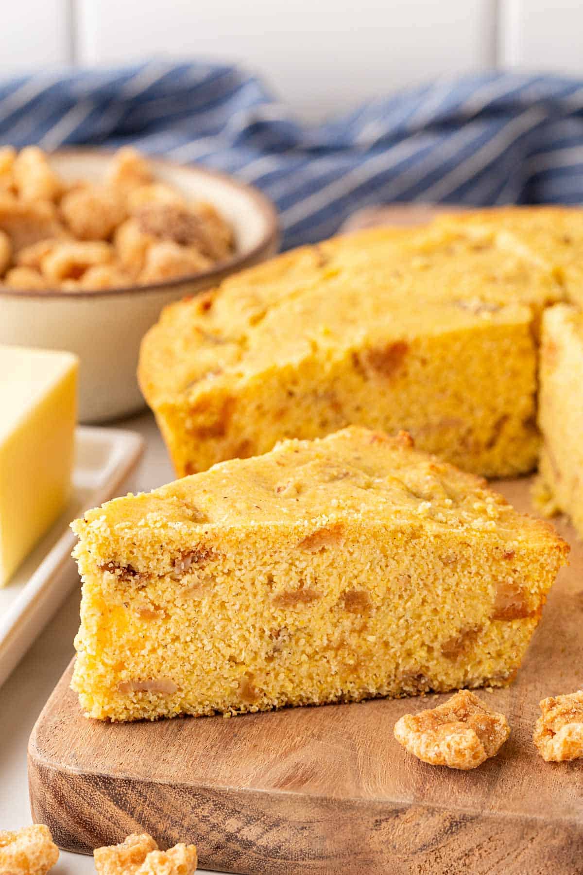 Thick slice of crackling bread on a wooden board with butter, pork cracklings, and a skillet of cornbread in the background
