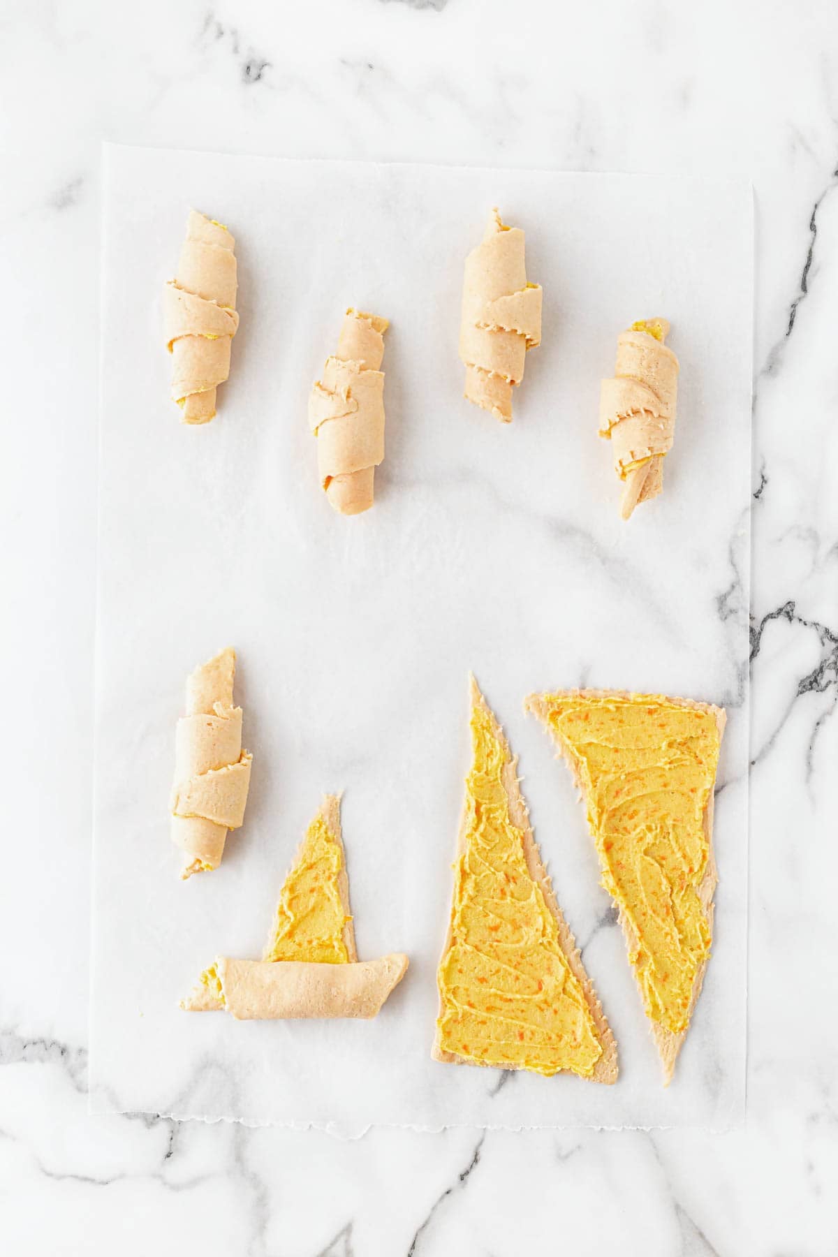 Bread dough triangles partially rolled into orange rolls on parchment paper over a white marble countertop
