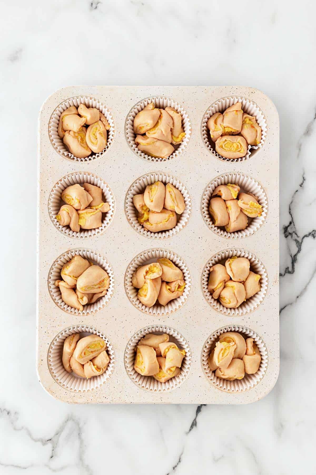 Orange roll pieces arranged in paper-lined muffin cups inside a muffin pan on a white marble countertop