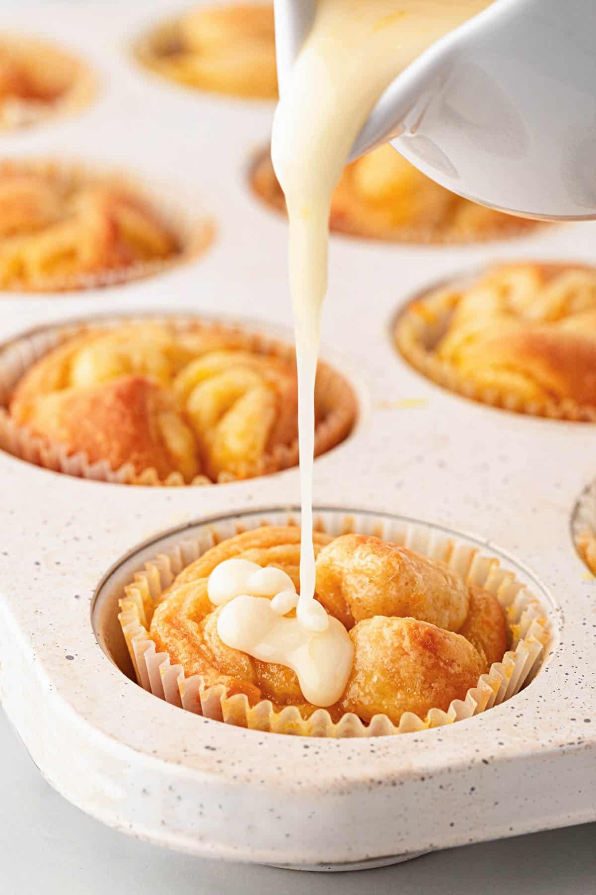 Orange glaze being poured over a warm orange roll in a muffin pan, with more baked rolls blurred in the background