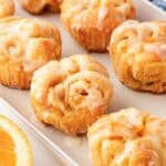 Multiple glazed orange rolls in a baking dish with fresh orange slices and a blue kitchen towel in the background