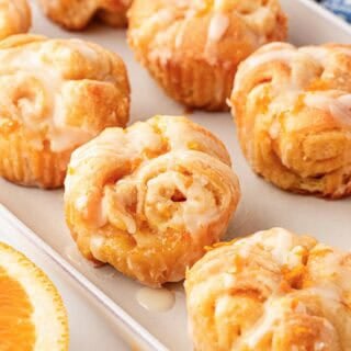 Multiple glazed orange rolls in a baking dish with fresh orange slices and a blue kitchen towel in the background