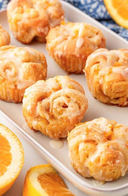 Multiple glazed orange rolls in a baking dish with fresh orange slices and a blue kitchen towel in the background