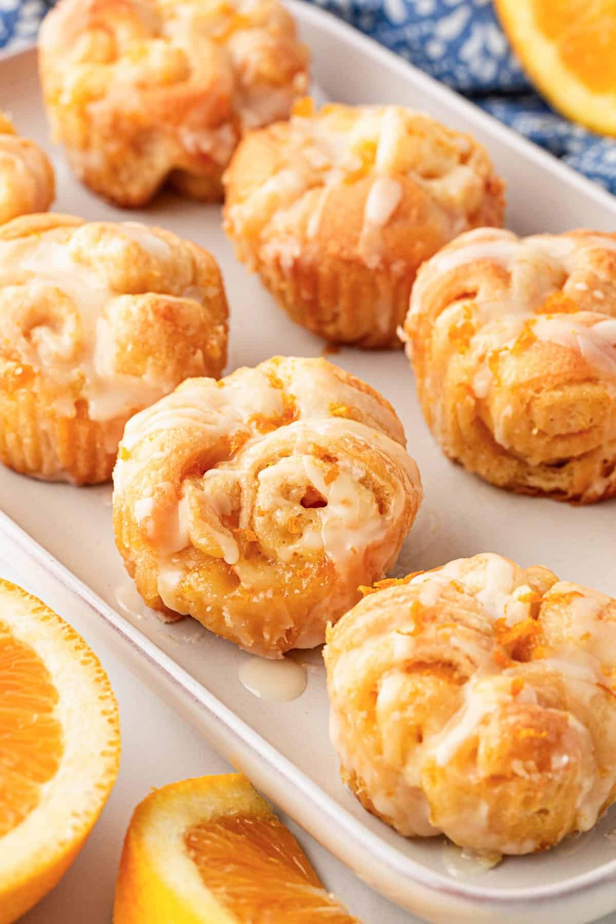 Multiple glazed orange rolls in a baking dish with fresh orange slices and a blue kitchen towel in the background
