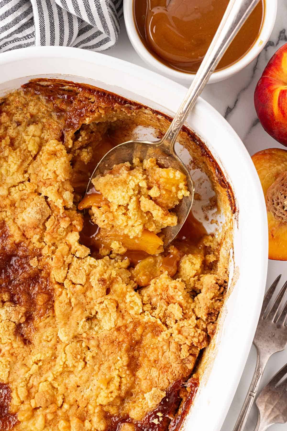 Spoon scooping peach dump cake from the baking dish with caramel sauce and fresh peaches nearby