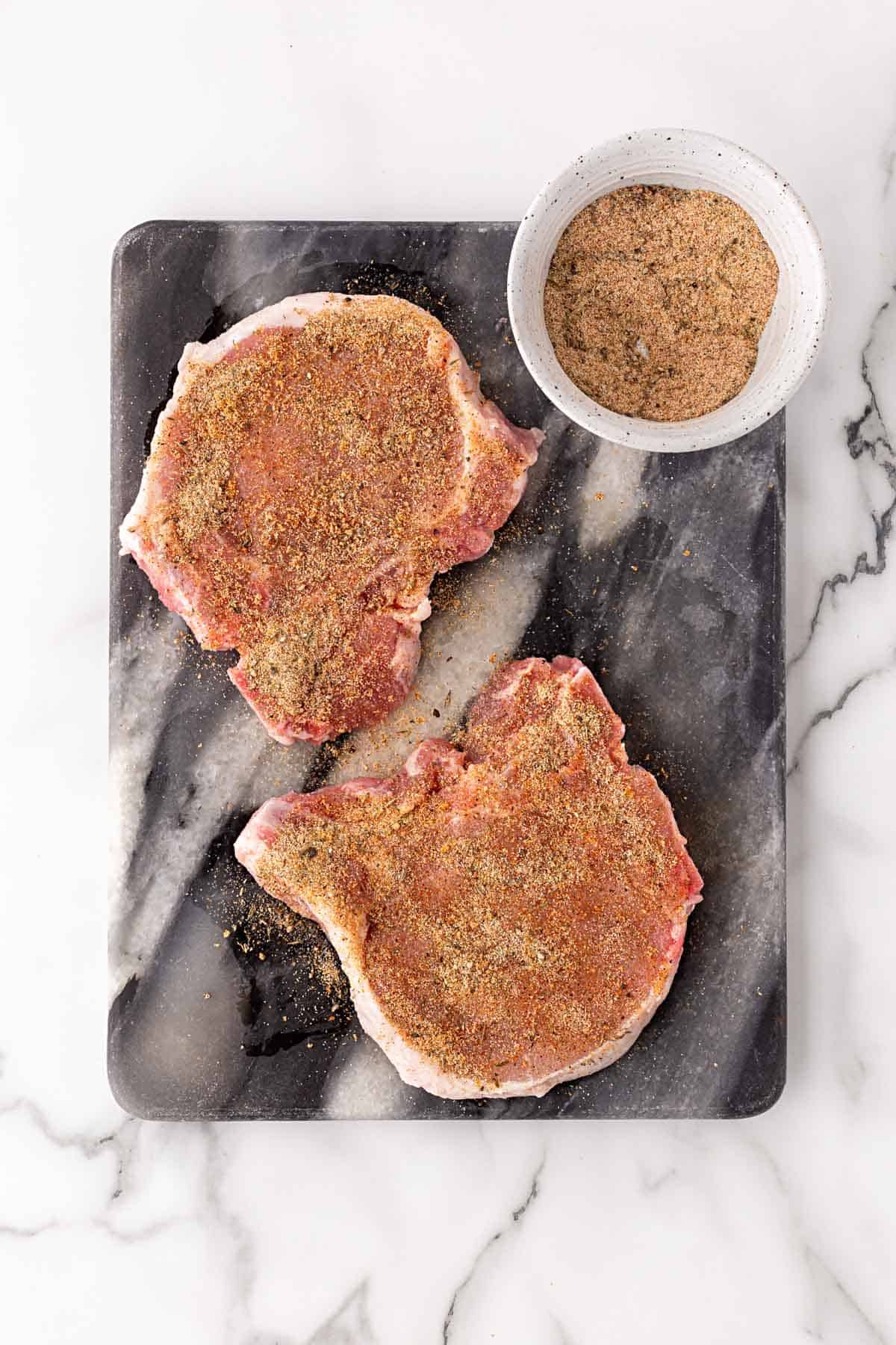 Two seasoned pork chops on a cutting board with a bowl of homemade seasoning