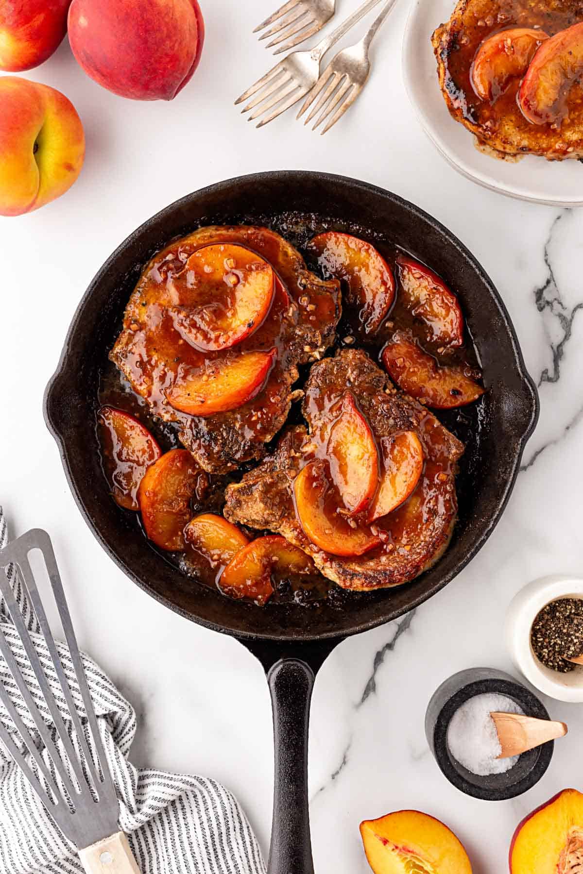 Cast iron skillet with peach pork chops topped with caramelized peaches and glaze, surrounded by fresh peaches and serving props