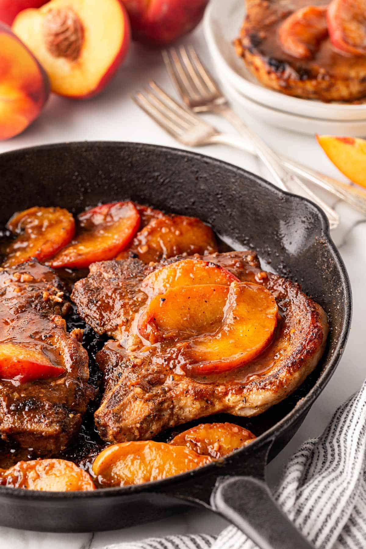 Peach pork chops in a cast iron skillet with fresh peaches, serving utensils, and a plated pork chop in the background
