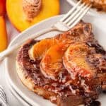 Closeup of a peach pork chop topped with caramelized peaches and glaze on a white plate, with fresh peaches and another plated pork chop in the background
