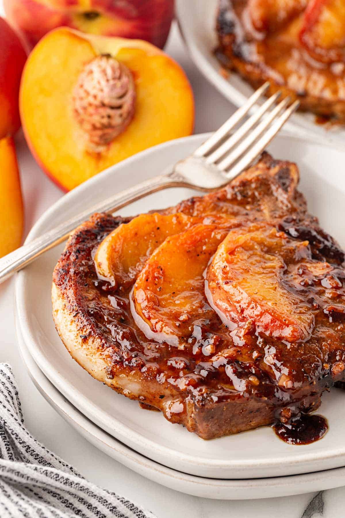 Closeup of a peach pork chop topped with caramelized peaches and glaze on a white plate, with fresh peaches and another plated pork chop in the background