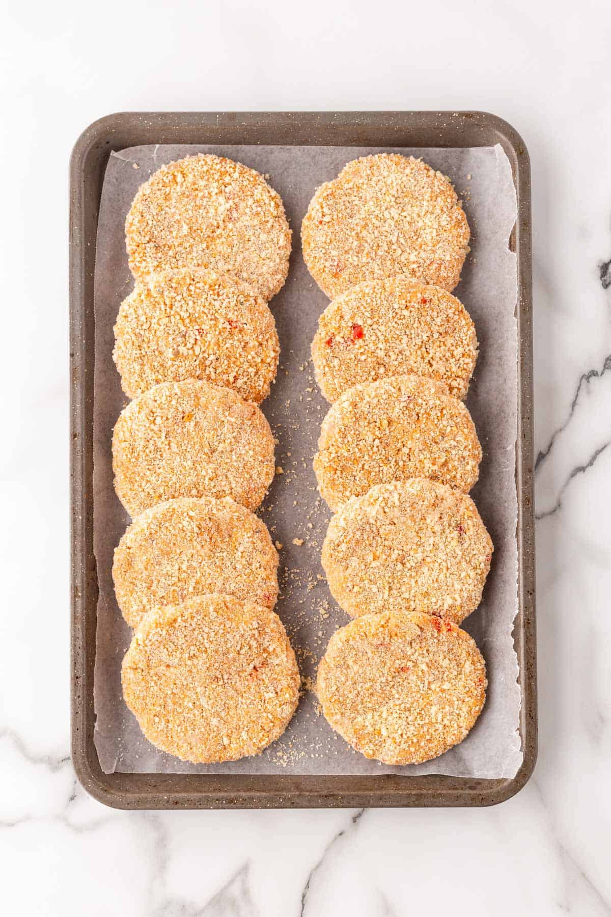 Breaded salmon cake patties on a parchment-lined baking sheet before frying on a marble countertop