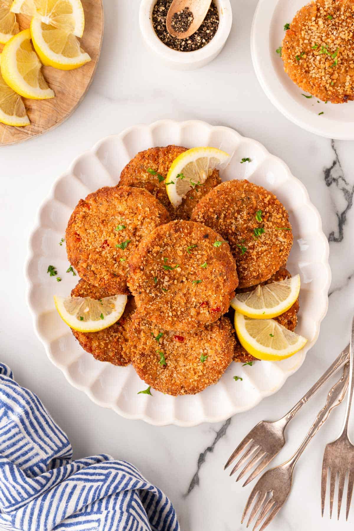 Platter of crispy salmon cakes garnished with parsley and lemon wedges on a marble countertop, with black pepper, extra salmon cakes, forks, and a blue striped kitchen towel in the background