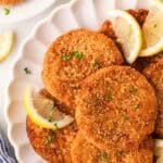 Overhead shot of crispy salmon cakes garnished with parsley and lemon wedges on a white serving platter, with black pepper and extra salmon cakes in the background