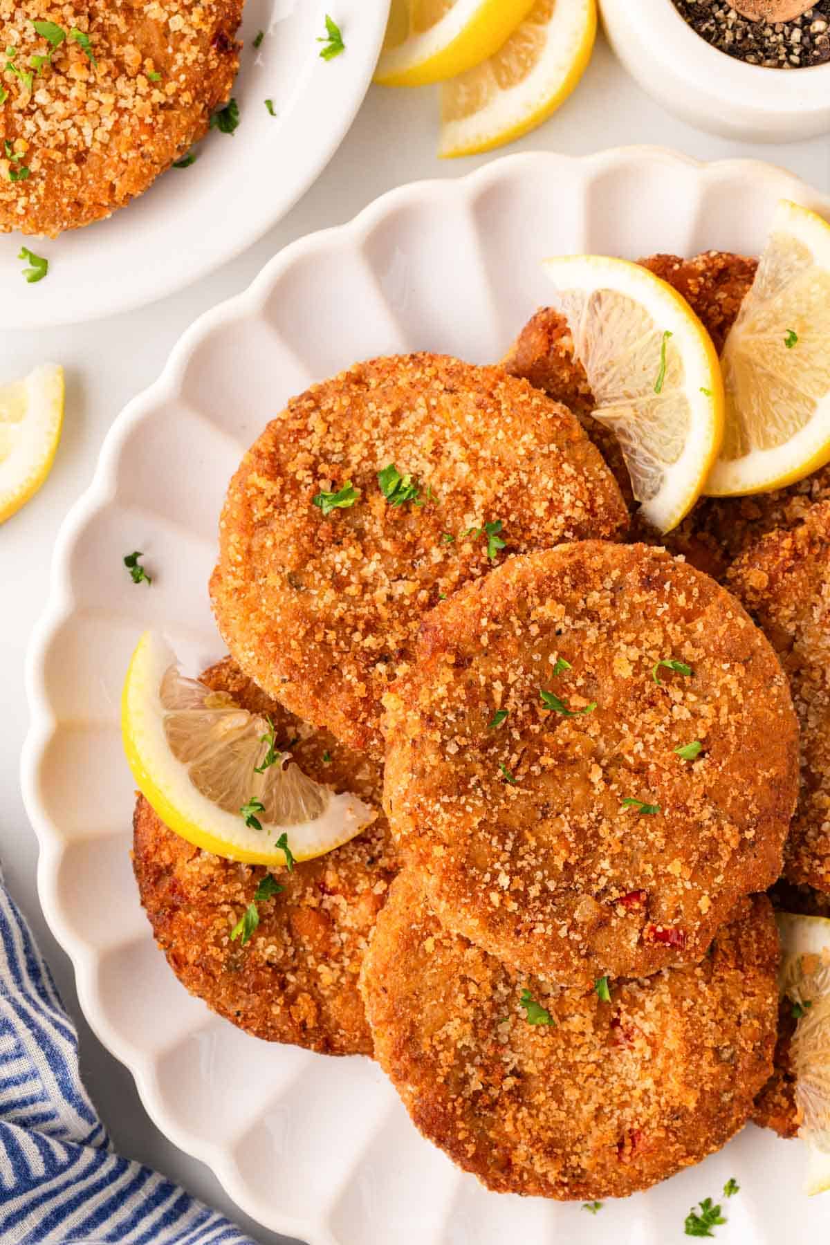 Overhead shot of crispy salmon cakes garnished with parsley and lemon wedges on a white serving platter, with black pepper and extra salmon cakes in the background