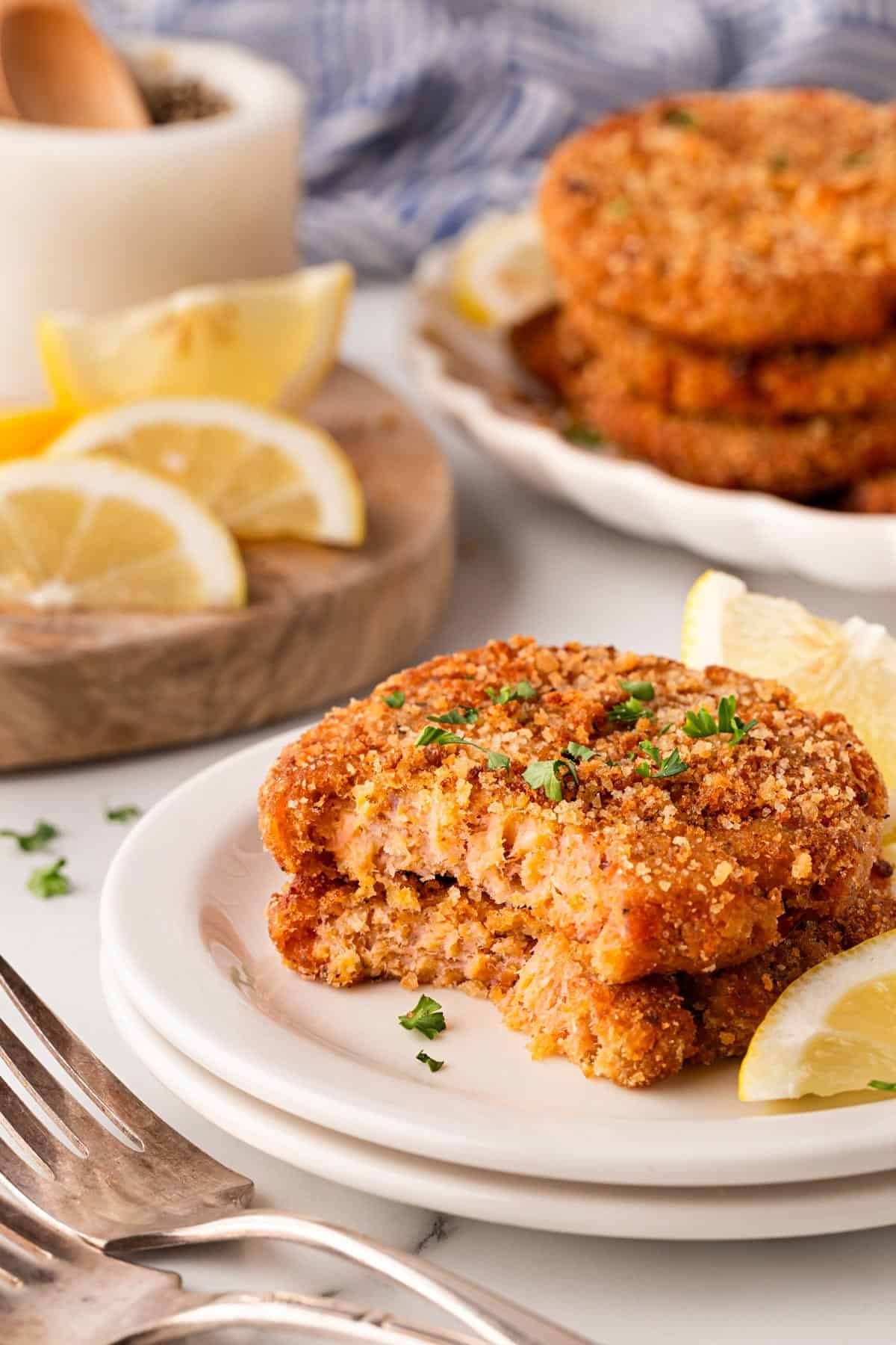 Salmon cakes broken open to show the flaky interior on a white plate, with lemon wedges and extra salmon cakes blurred in the background