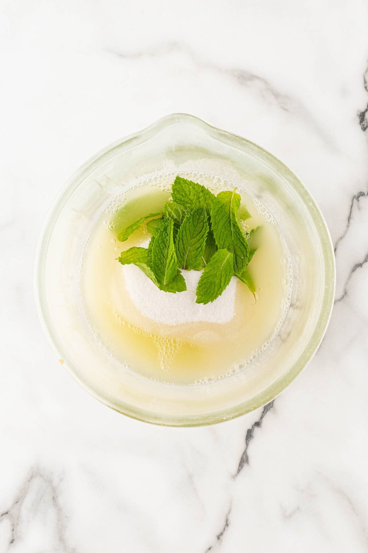 Bowl with lemon juice, sugar, and fresh mint leaves on a white marble countertop before muddling