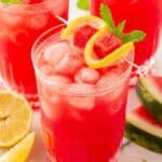 Three glasses of watermelon lemonade garnished with lemon slices, watermelon cubes, and fresh mint on a marble countertop