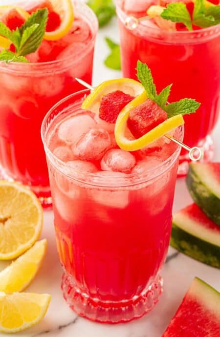 Three glasses of watermelon lemonade garnished with lemon slices, watermelon cubes, and fresh mint on a marble countertop