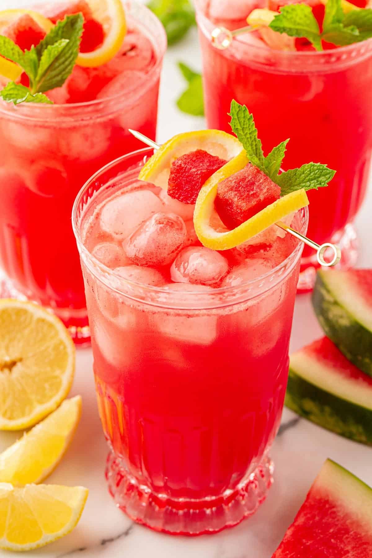 Three glasses of watermelon lemonade garnished with lemon slices, watermelon cubes, and fresh mint on a marble countertop