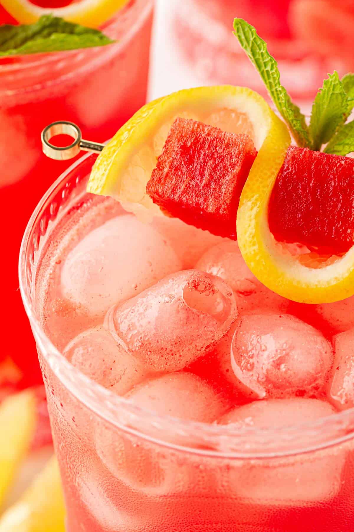 Closeup of watermelon lemonade topped with ice, lemon slices, watermelon cubes, and mint, with another glass softly blurred behind it