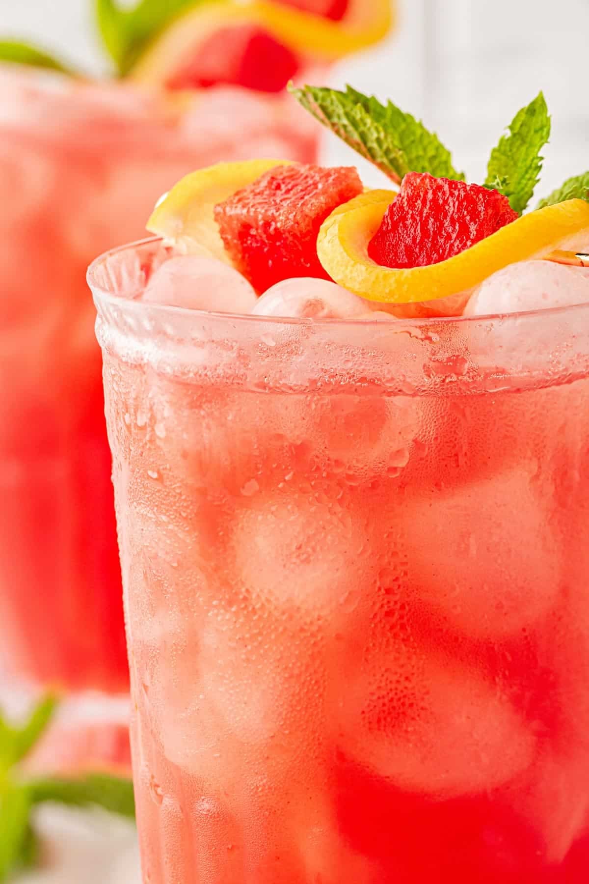 Closeup of a glass of watermelon lemonade with ice, mint, lemon slices, and fresh watermelon cubes, with more drinks blurred in the background