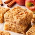 Closeup of stacked apple pie bars with fresh apples and cinnamon sticks in the background