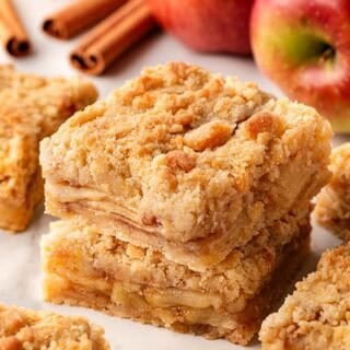 Closeup of stacked apple pie bars with fresh apples and cinnamon sticks in the background