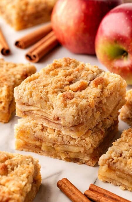 Closeup of stacked apple pie bars with fresh apples and cinnamon sticks in the background