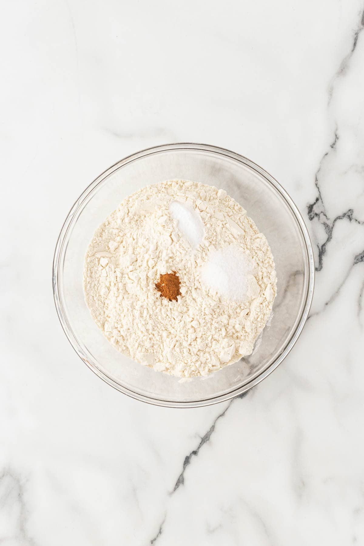 Flour, baking soda, salt, and cinnamon in a glass mixing bowl on a white marble surface
