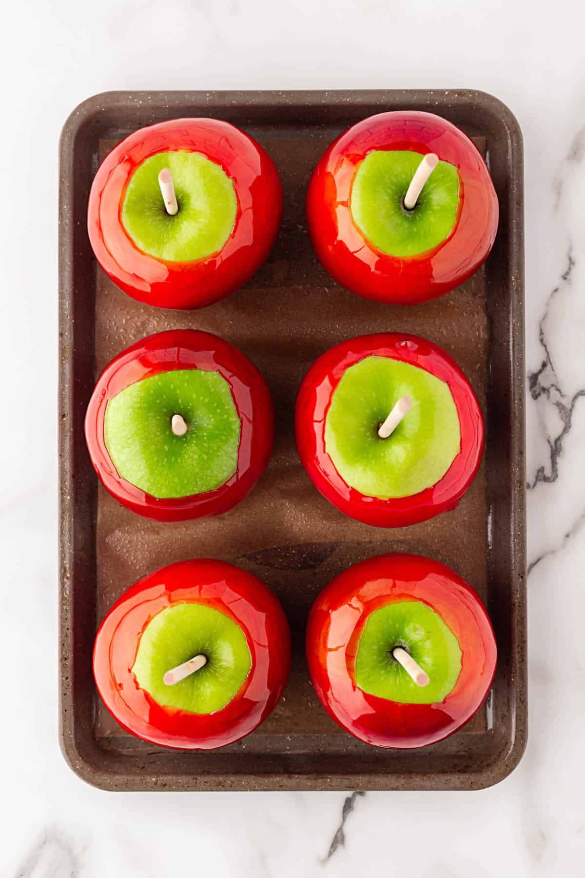 Six freshly coated red candy apples resting on a baking sheet