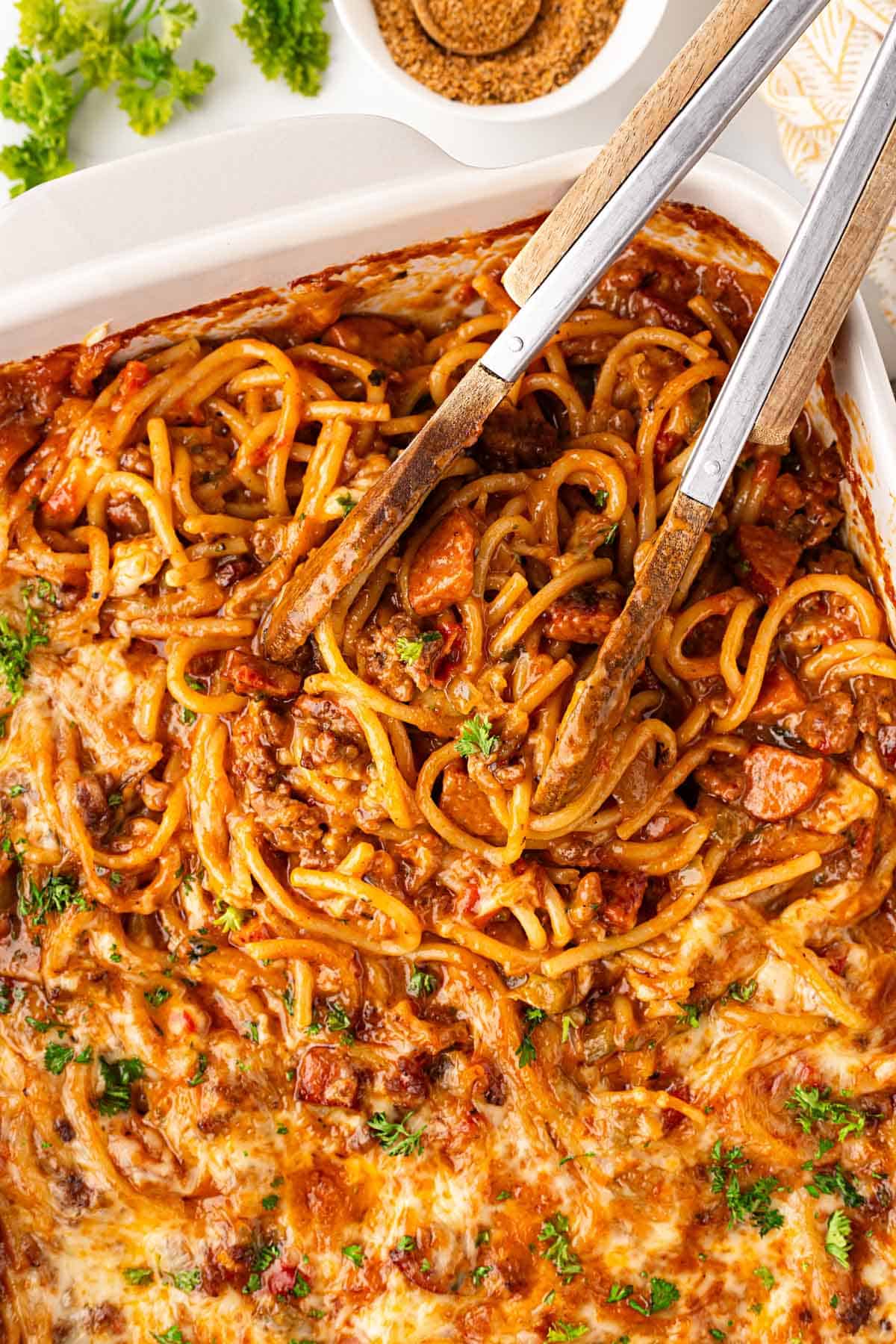 Overhead shot of cheesy baked dirty spaghetti with beef and andouille sausage being lifted with serving tongs