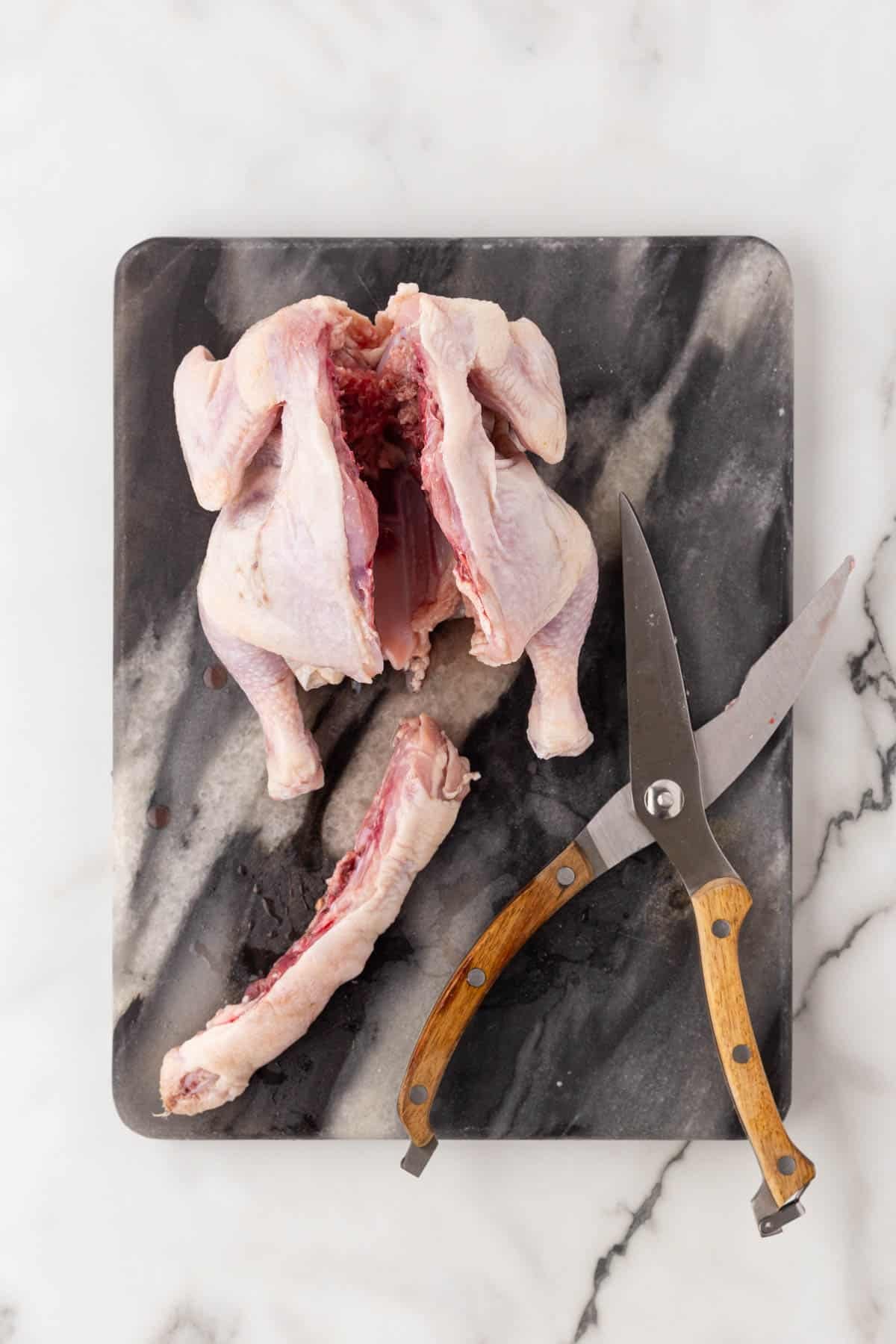 Raw Cornish hen with the backbone removed, alongside kitchen shears and the removed backbone on a dark marble cutting board