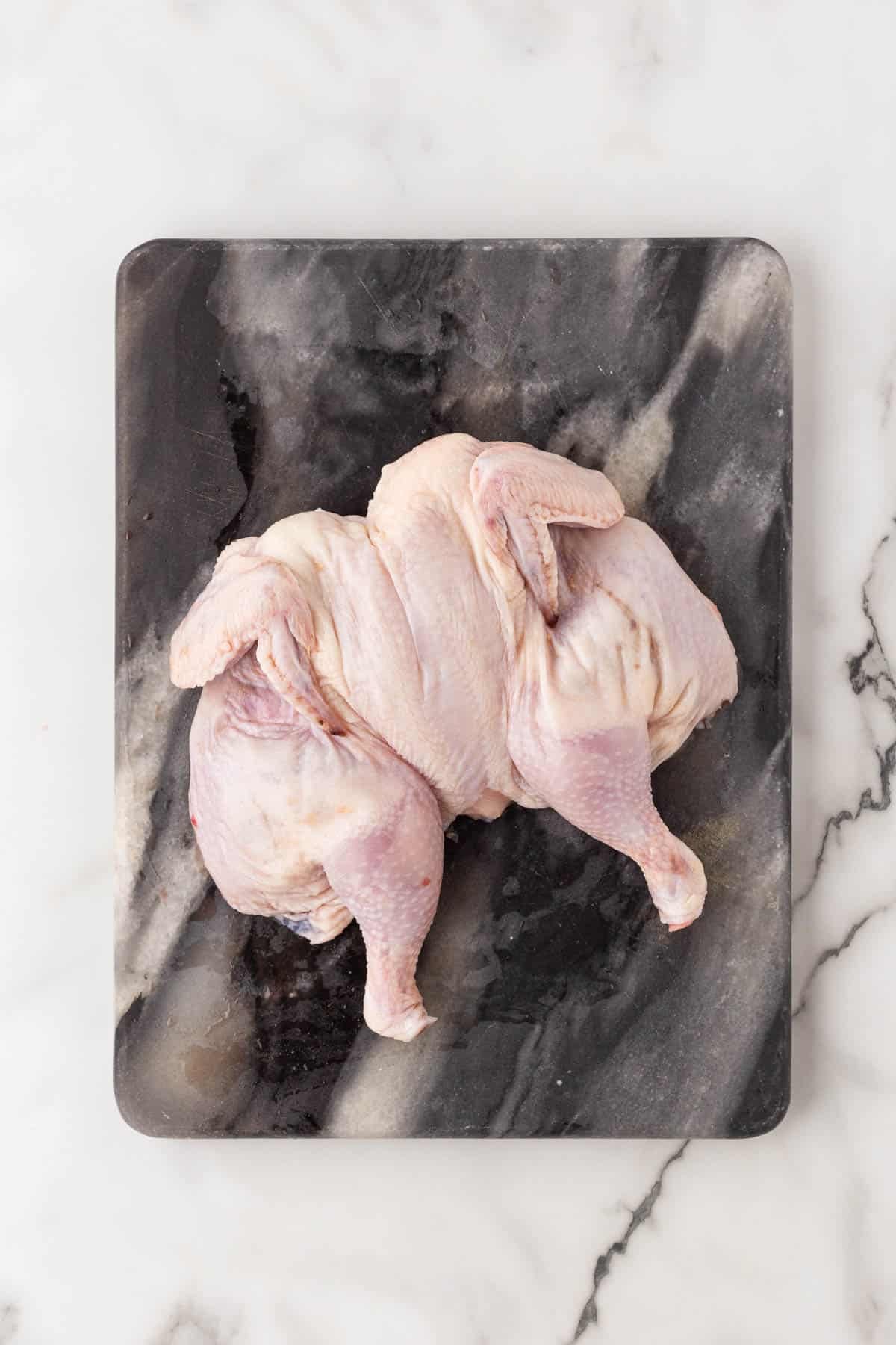 Raw spatchcocked Cornish hen flattened breast-side up on a dark marble cutting board
