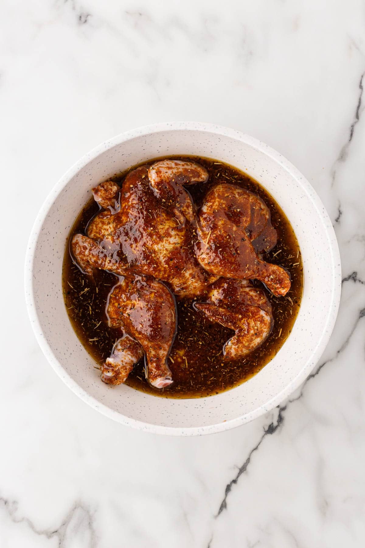 Raw Cornish hens coated in a dark, seasoned marinade inside a large white bowl on a marble countertop