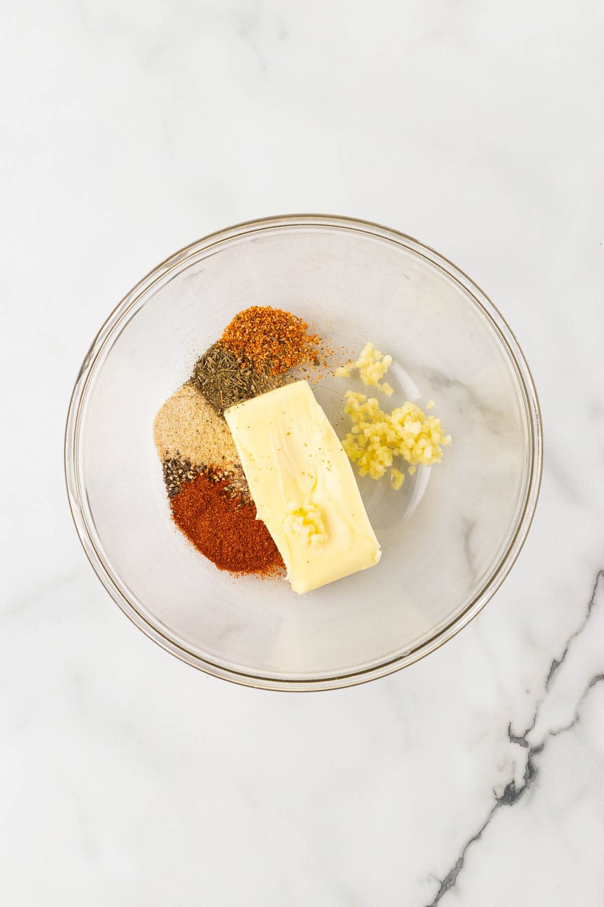 Softened butter with minced garlic and assorted spices in a clear glass bowl on a white marble countertop