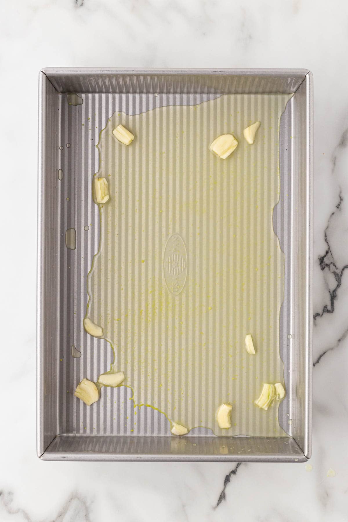 Metal baking pan with chicken broth and smashed garlic cloves spread across the bottom