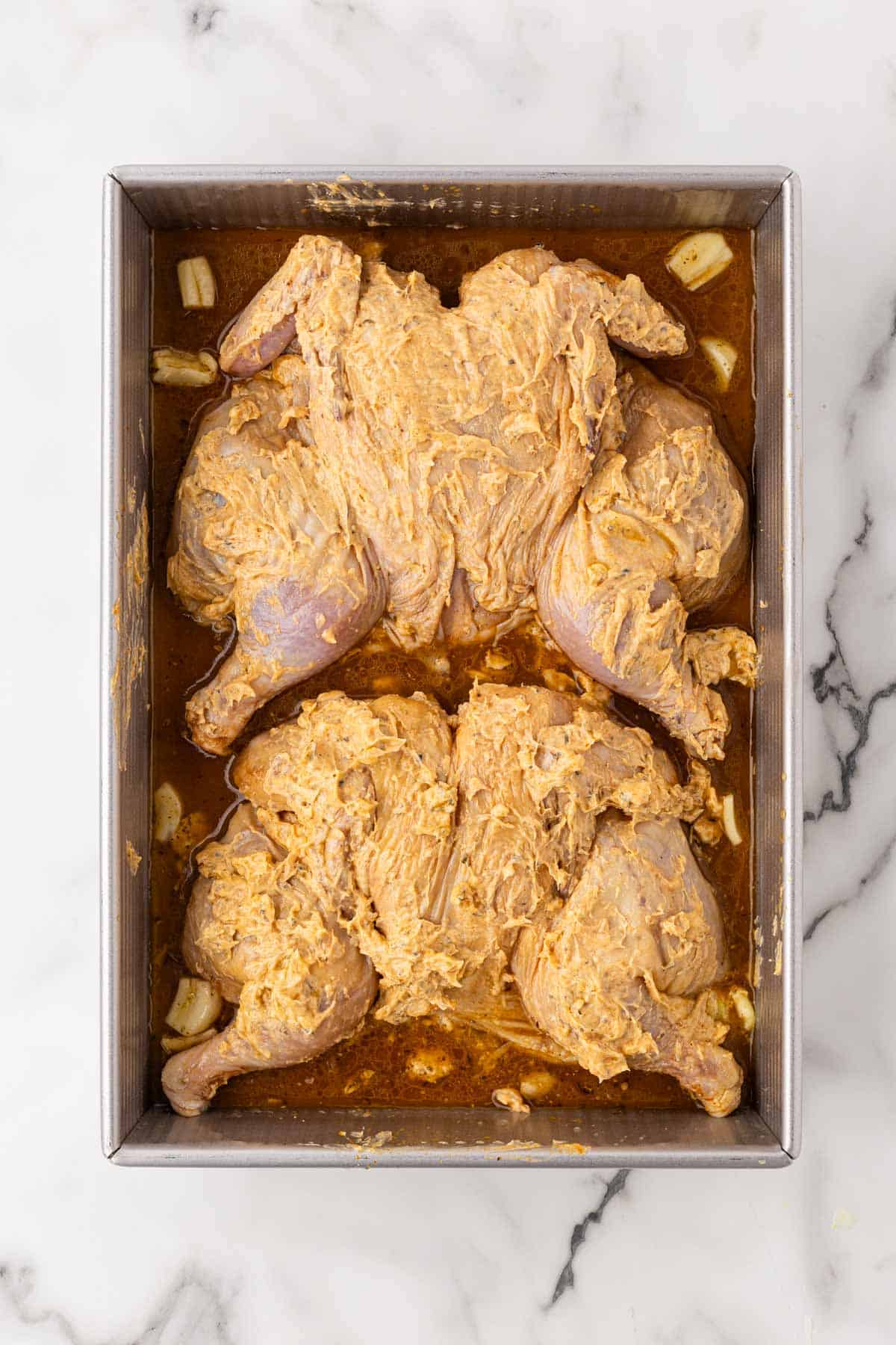Two raw spatchcocked Cornish hens coated in seasoned compound butter inside a metal baking pan with broth and garlic