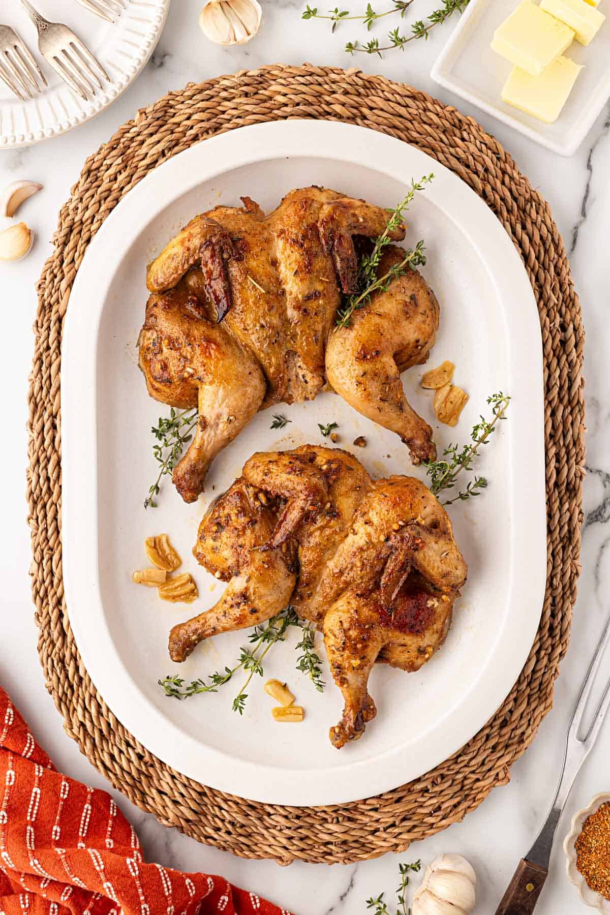 Two roasted Cornish hens with golden, seasoned skin on a white serving platter garnished with fresh thyme and garlic
