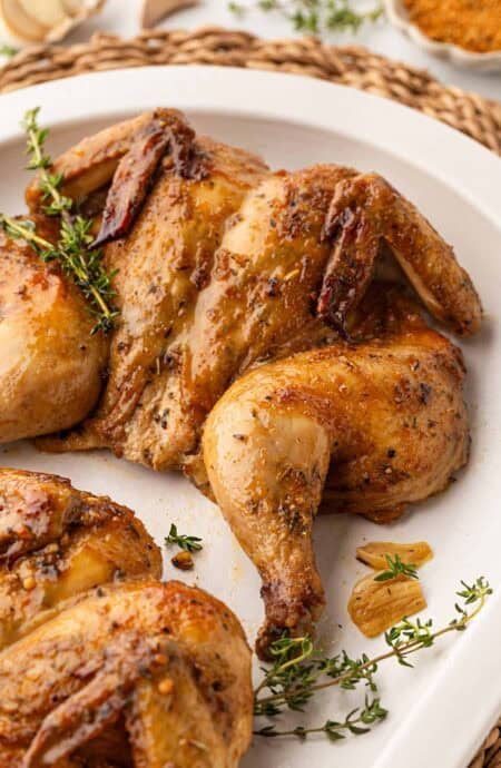 Closeup of a roasted Cornish hen with crispy, seasoned skin, surrounded by fresh thyme and roasted garlic on a white platter