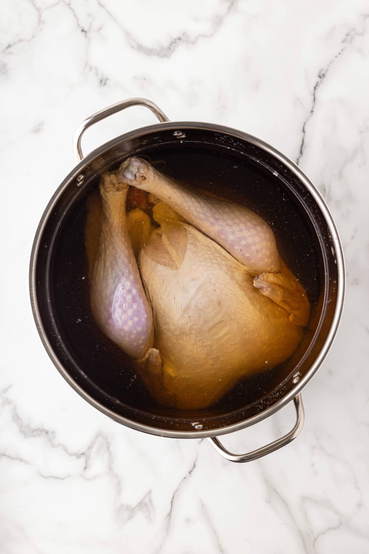 Whole raw turkey submerged in brine inside a large stainless steel pot on a marble countertop