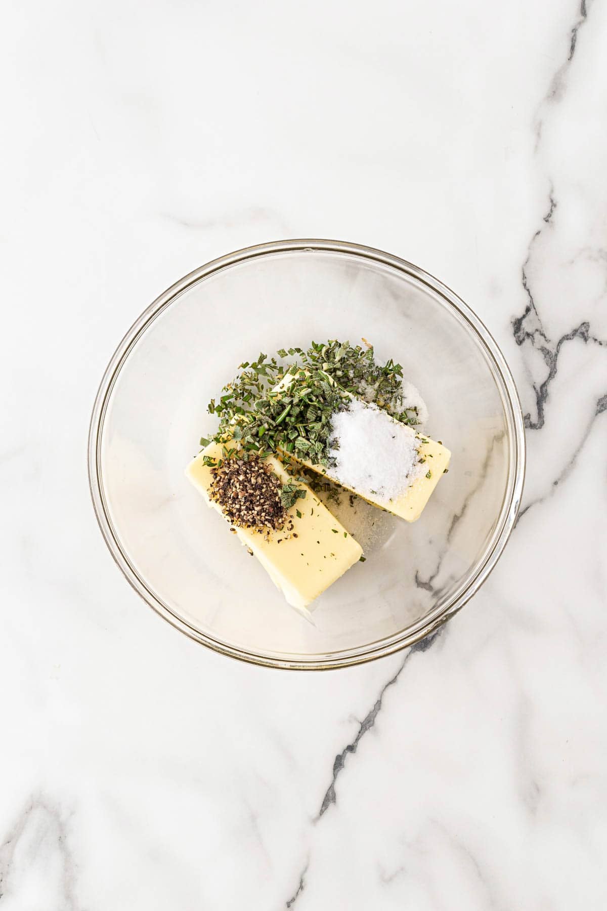 Softened butter topped with chopped fresh herbs, kosher salt, and black pepper in a clear glass bowl on a marble countertop