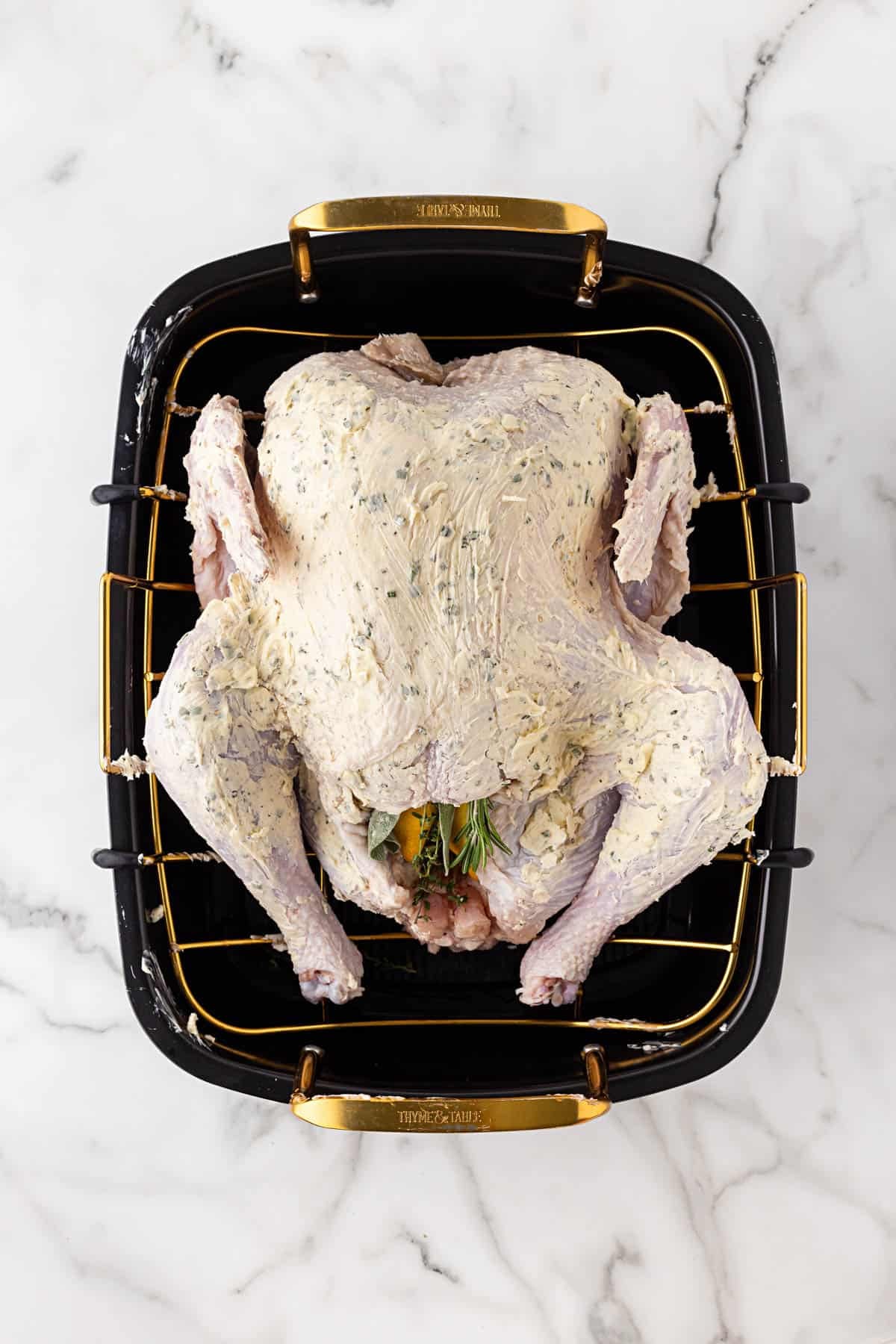 Raw turkey coated in herb butter with fresh herbs and lemon in the cavity, sitting on a gold roasting rack in a black pan
