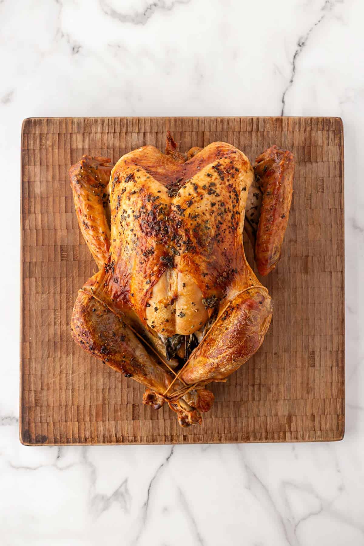 Whole roasted turkey with golden-brown skin and trussed legs resting on a wooden cutting board over a marble countertop
