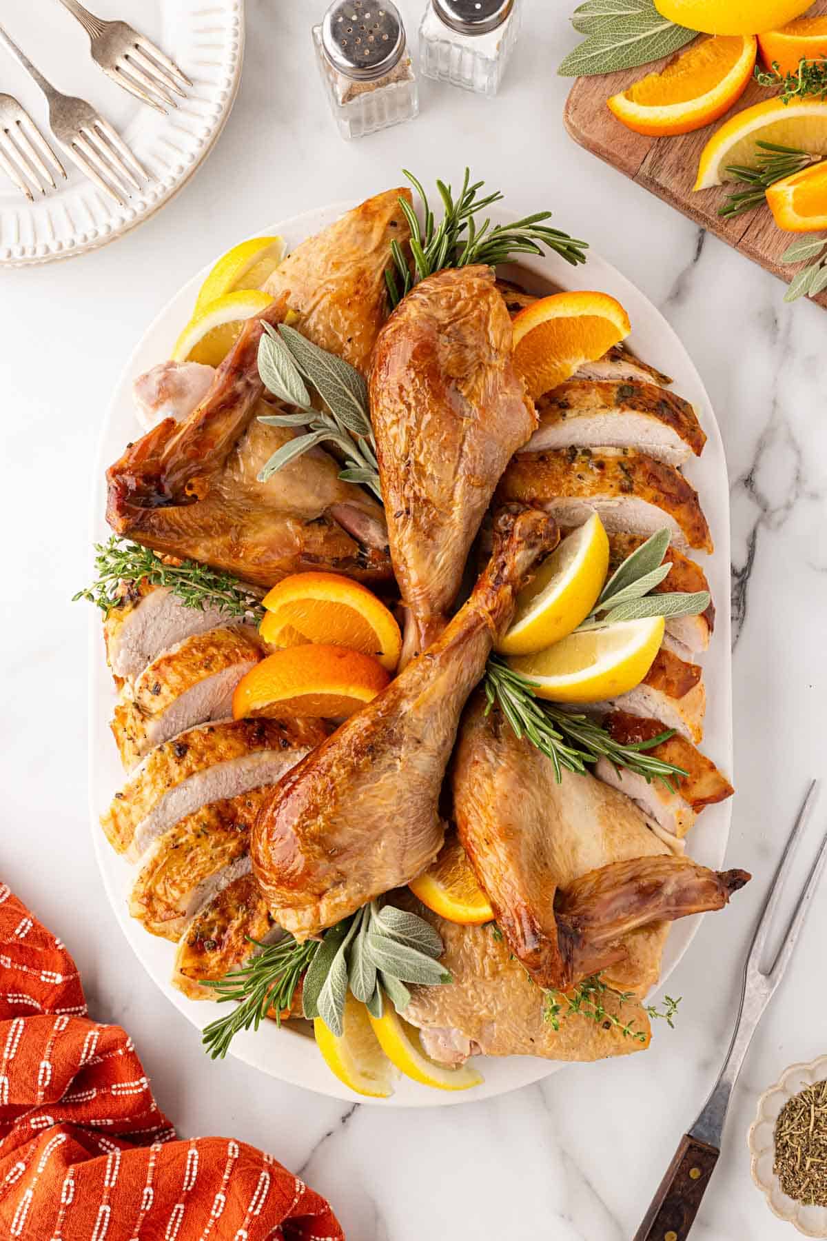 Carved oven roasted turkey arranged on a white platter with fresh rosemary, sage, thyme, lemon wedges, and orange slices