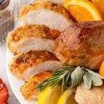 Closeup of sliced oven roasted turkey breast with golden skin, a roasted drumstick, fresh herbs, and citrus wedges on a white platter