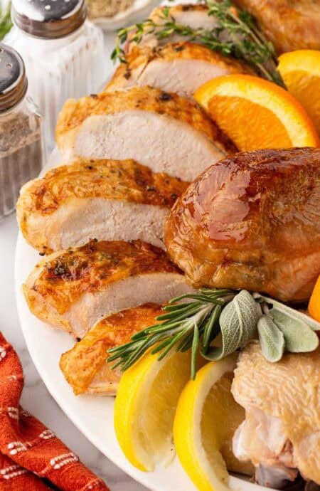 Closeup of sliced oven roasted turkey breast with golden skin, a roasted drumstick, fresh herbs, and citrus wedges on a white platter