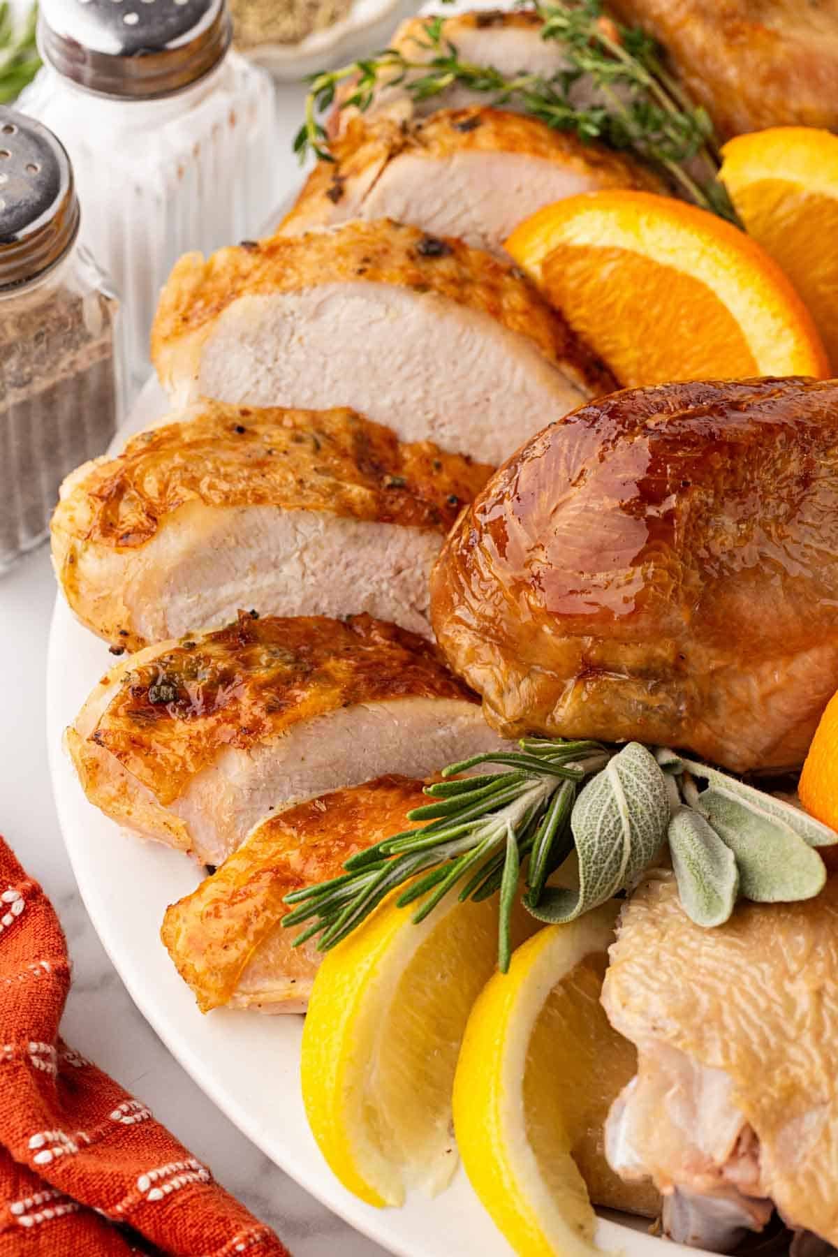 Closeup of sliced oven roasted turkey breast with golden skin, a roasted drumstick, fresh herbs, and citrus wedges on a white platter