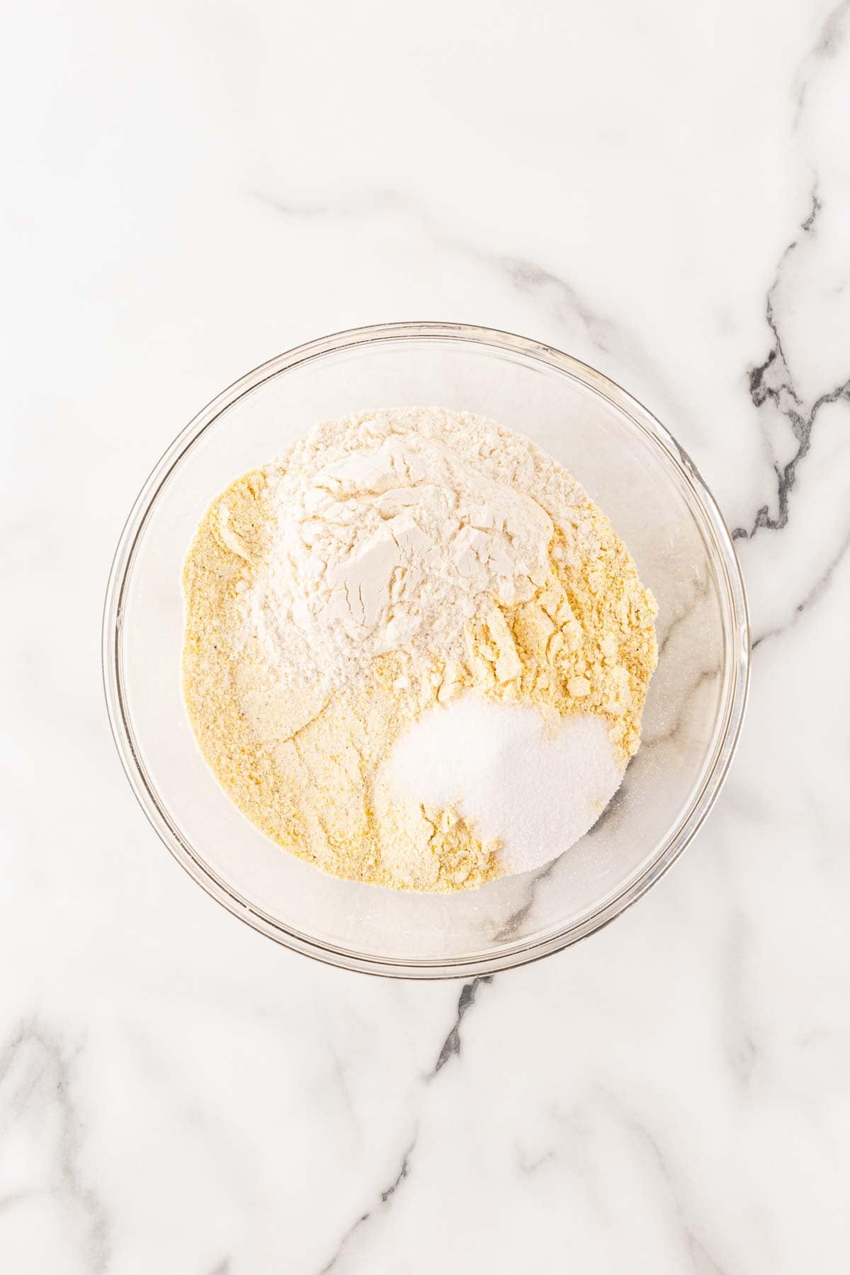 Self-rising cornmeal, flour, and sugar in a clear glass mixing bowl on a white marble countertop