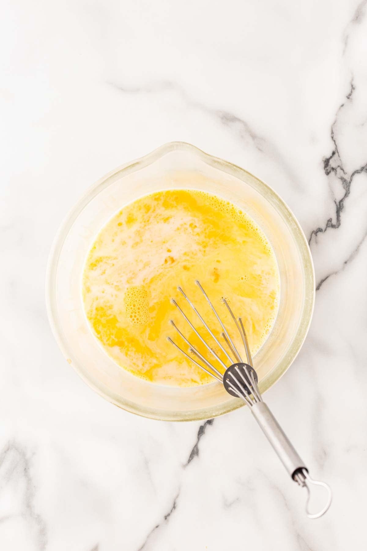 Milk and beaten eggs mixed in a glass bowl with a metal whisk on a marble countertop
